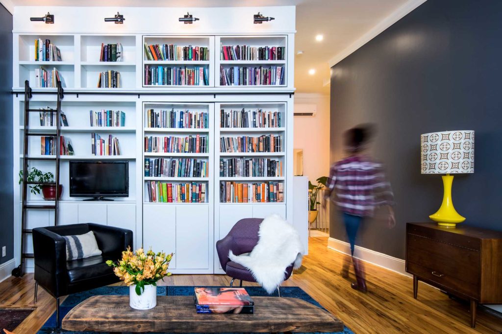 Hiring for your design project: white custom shelving with books and ladder in a modern living room with dark blue walls, wood floor and area rug; a woman walks down the hall away from the camera.