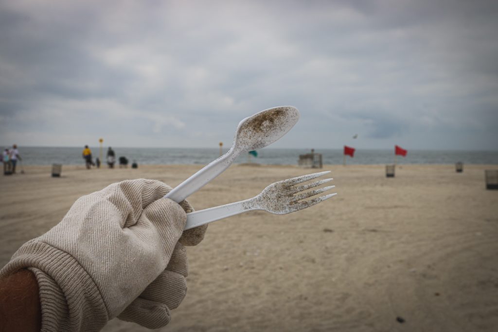 Dirty plastic cutlery held up by a gloved hand on an overcast beach with beachgoers in the background.