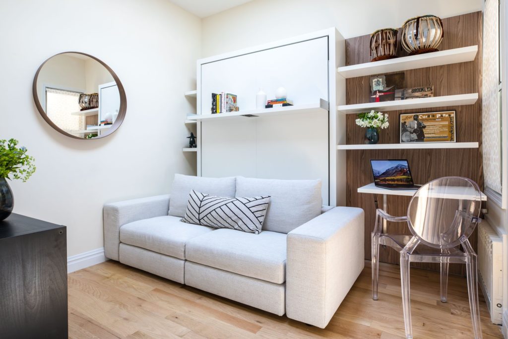Creating a space plan: home office with wood floor, walnut and white shelving, lucite chair, circular mirror and grey sofa in front of closed Murphy bed.