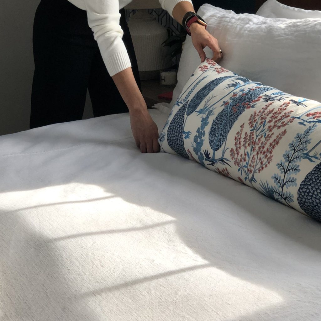 Designing based on someone else's preferences: a woman's hands adjust a blue, white and red plant themed lumbar pillow on a white linen bedspread.
