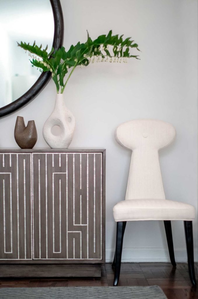 MInimalism and maximalism: white, modern chair next to brown cabinet with white maze graphic, organic vases and round mirror. 