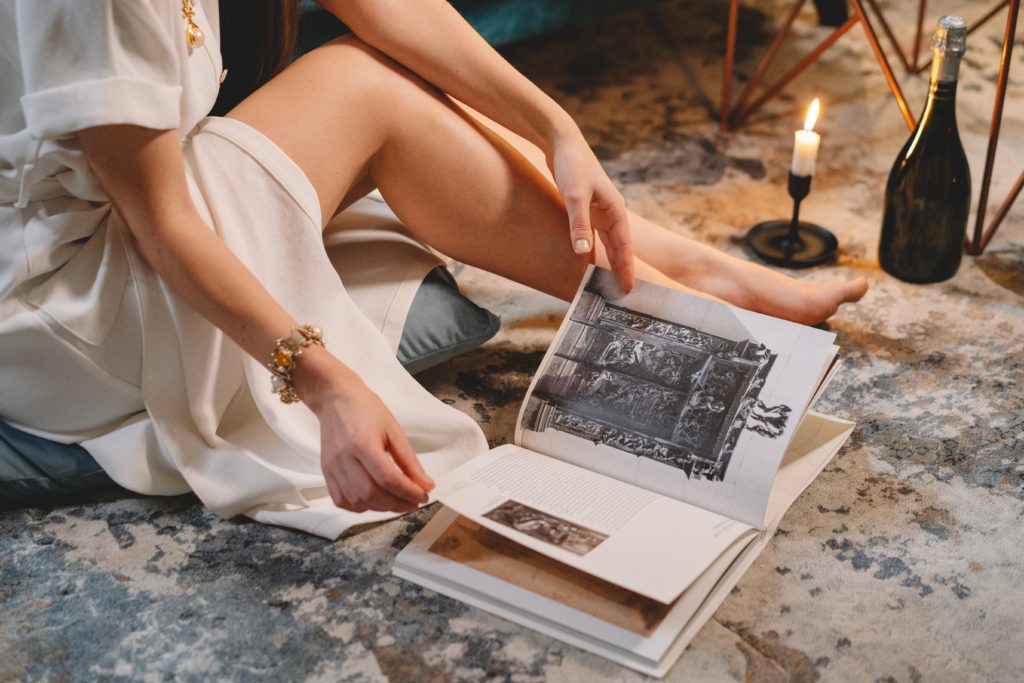 Woman in white dress reading art book on luxury rug with candle and wine.