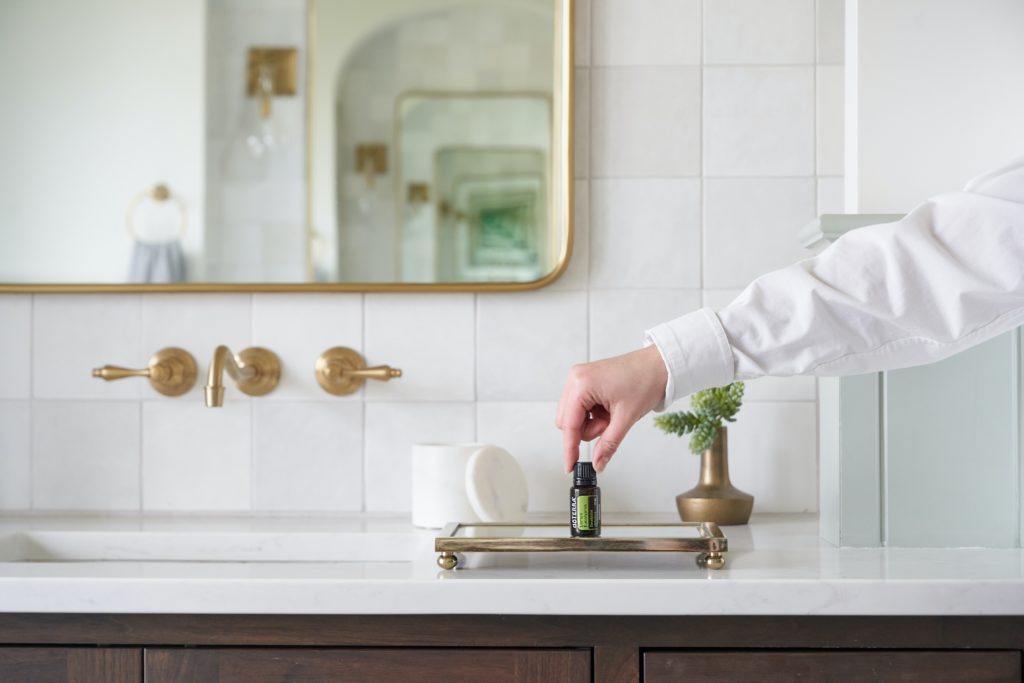 Final touches in your design project: white marble bathroom with brass fixtures, and hand placing essential oil bottle on tray.
