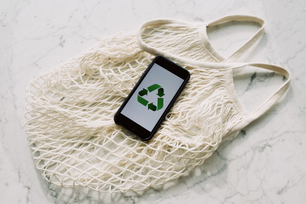 Reusable mesh shopping bag with smartphone sitting on top, displaying the reduce, reuse, recycle symbol.