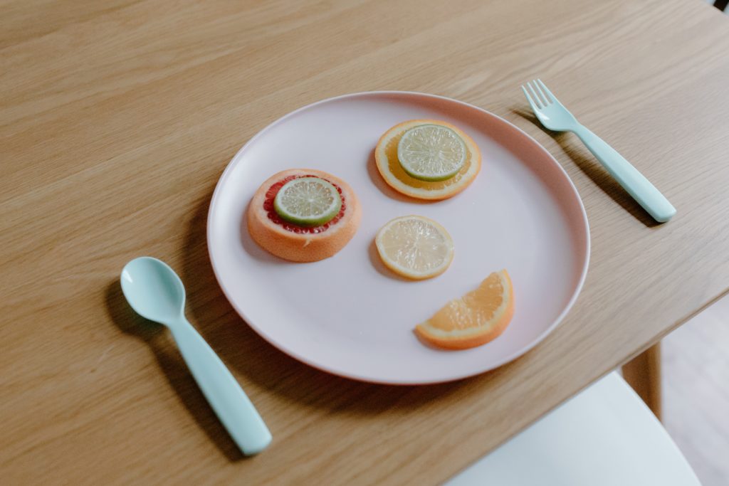 Pink plastic plate and cutlery with citrus slices coming a smile, all sitting on a wood tabletop.