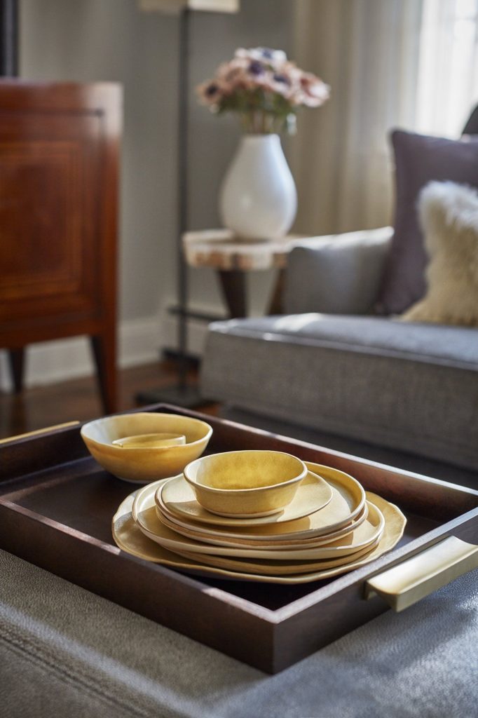 In terms of design: yellow, organic shaped ceramic ware on a wooden tray sits on a grey ottoman with grey sofa in background.