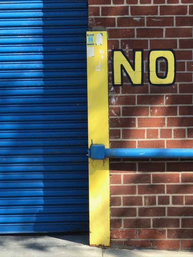 The psychology of hidden desire: a brick wall with a bright blue garage door, and a yellow painted "no" over the brick.