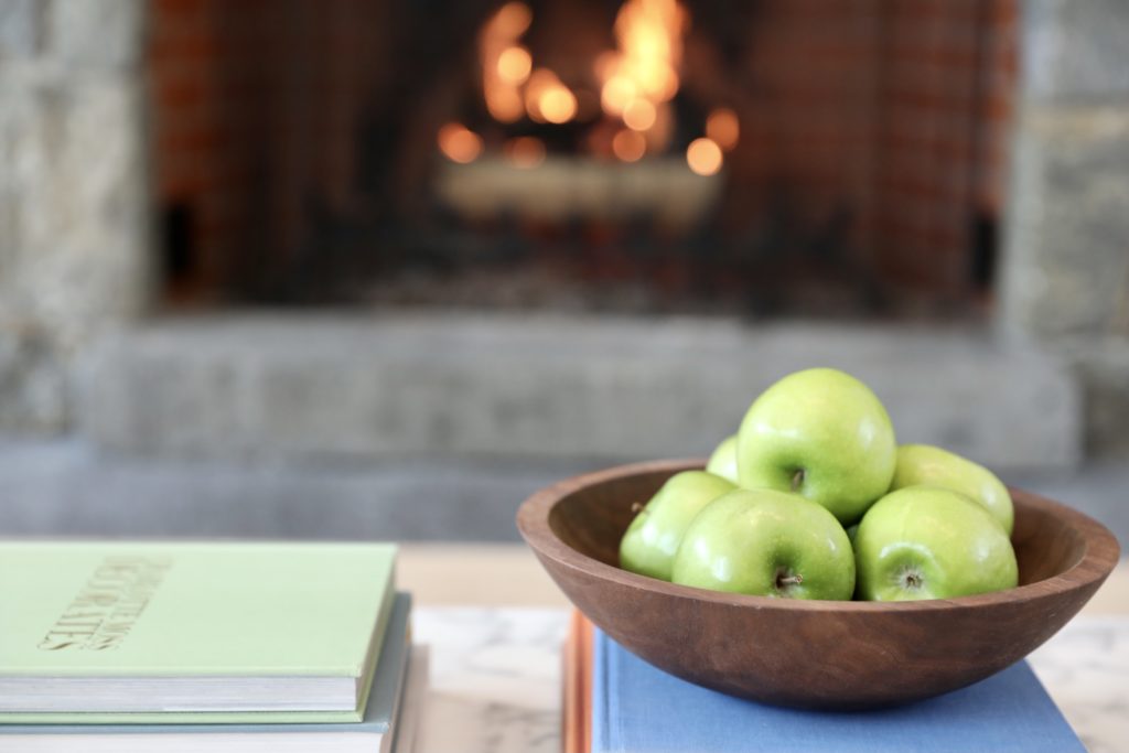 Walnut bowl of Granny Smith apples on stack of books, with fireplace in background.