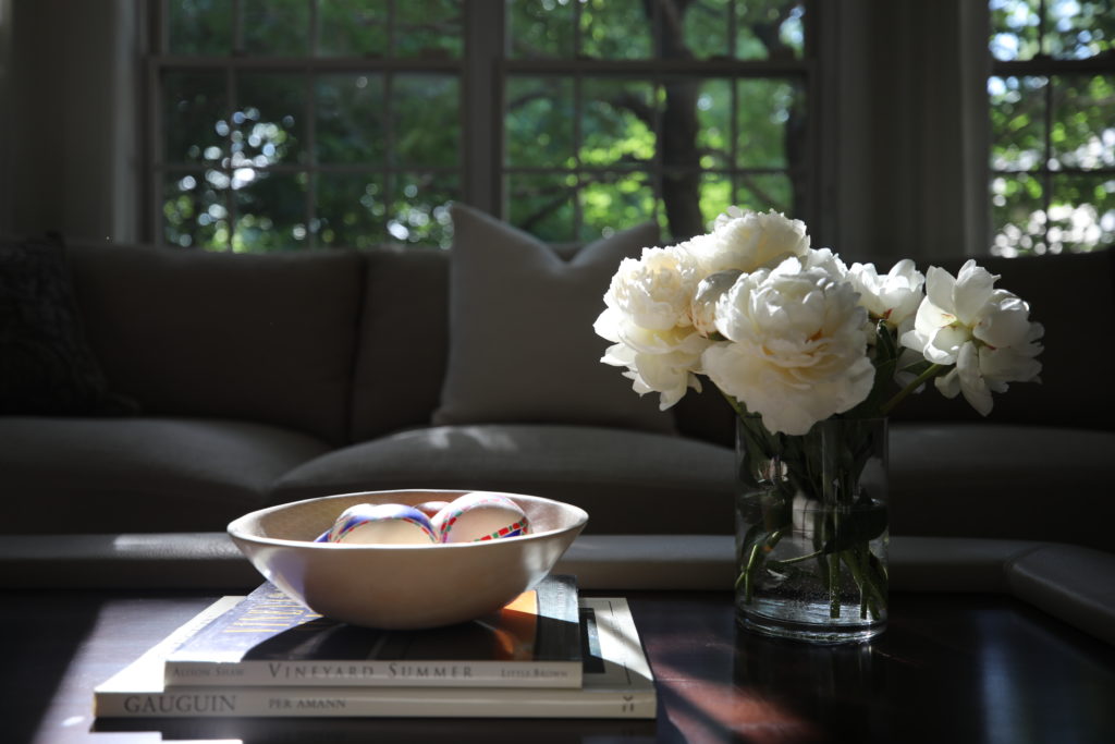 Working with an interior designer: late afternoon light piercing a living room with walnut coffee table, white blooms and grey sofa in background.