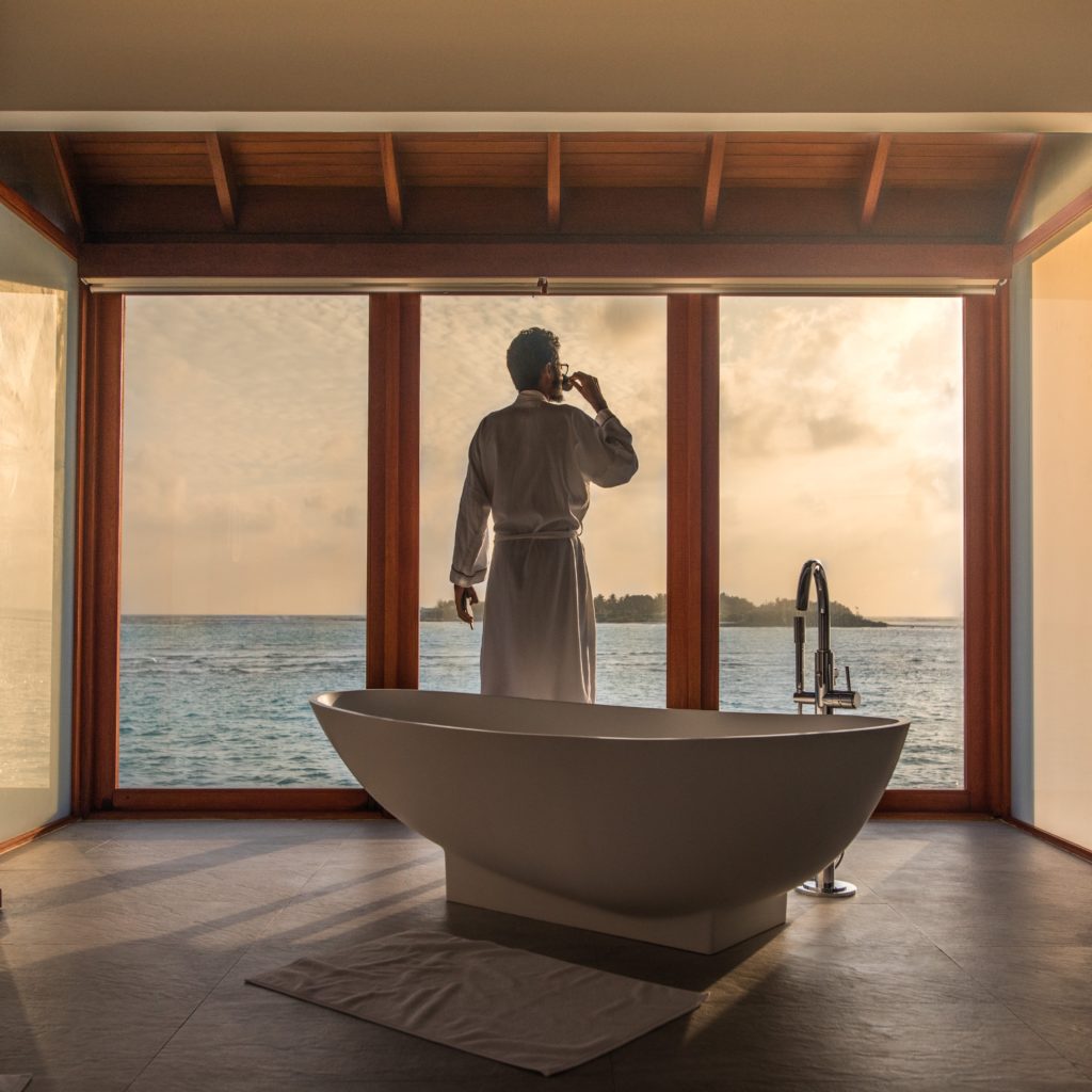 Self-actualization through interior design: man in robe drinks coffee in front of window featuring sunset ocean view, with modern tub in foreground.