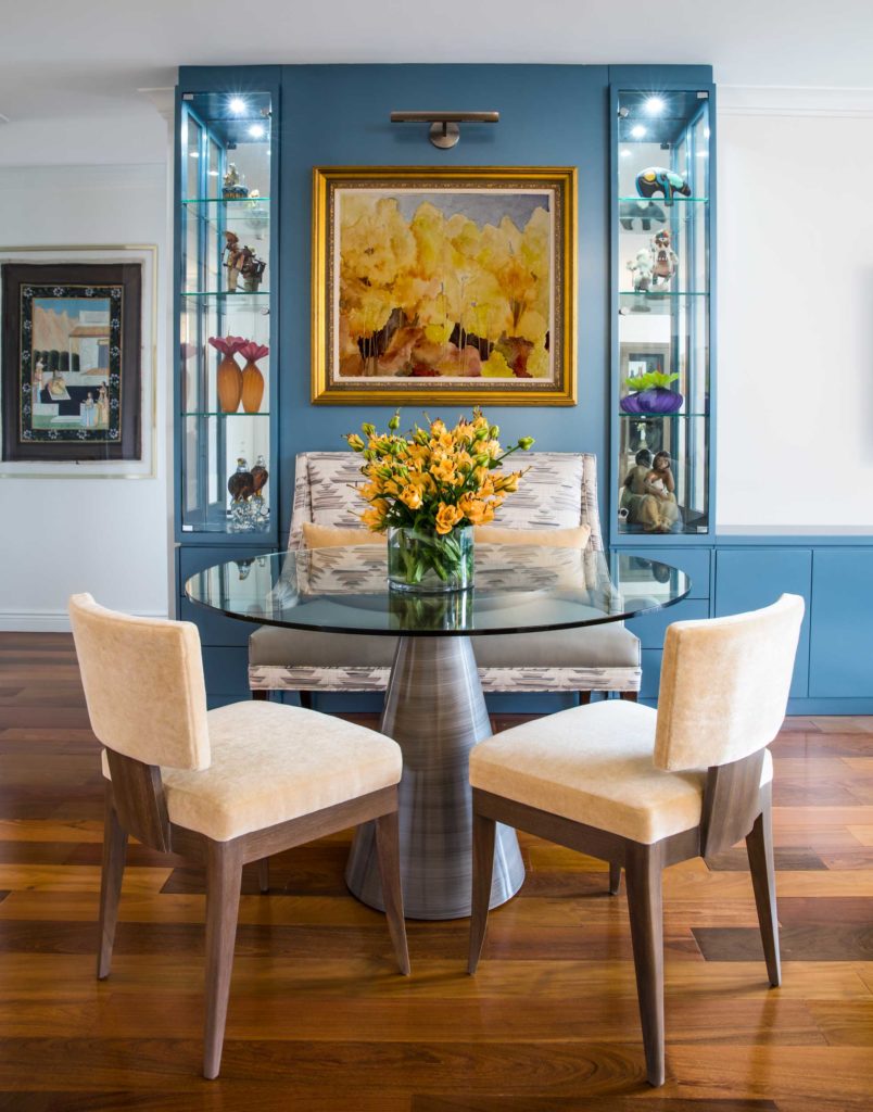 Playing the long game with luxury: blue, white and wood dining room with yellow flower painting, yellow flowers, glass top dining table and glass display cases with vases.