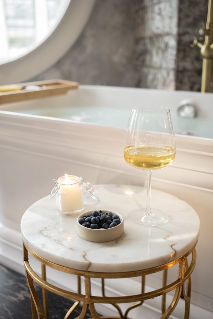 Self-actualization through interior design: marble and brass side table with wine, candle and blueberries foreground a full white porcelain tub in the background.