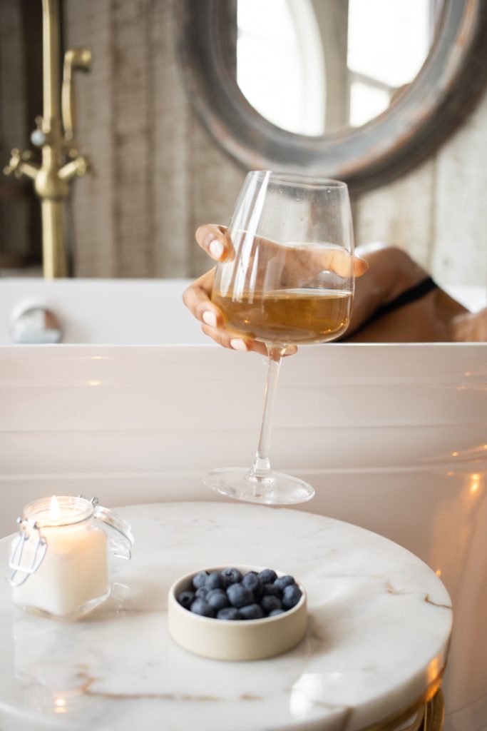 Self Actualization: a woman's hand emerges from a white bathtub, holding a glass of white wine, suspended over a marble side table with blueberries in a dish and a white candle in a small mason jar.