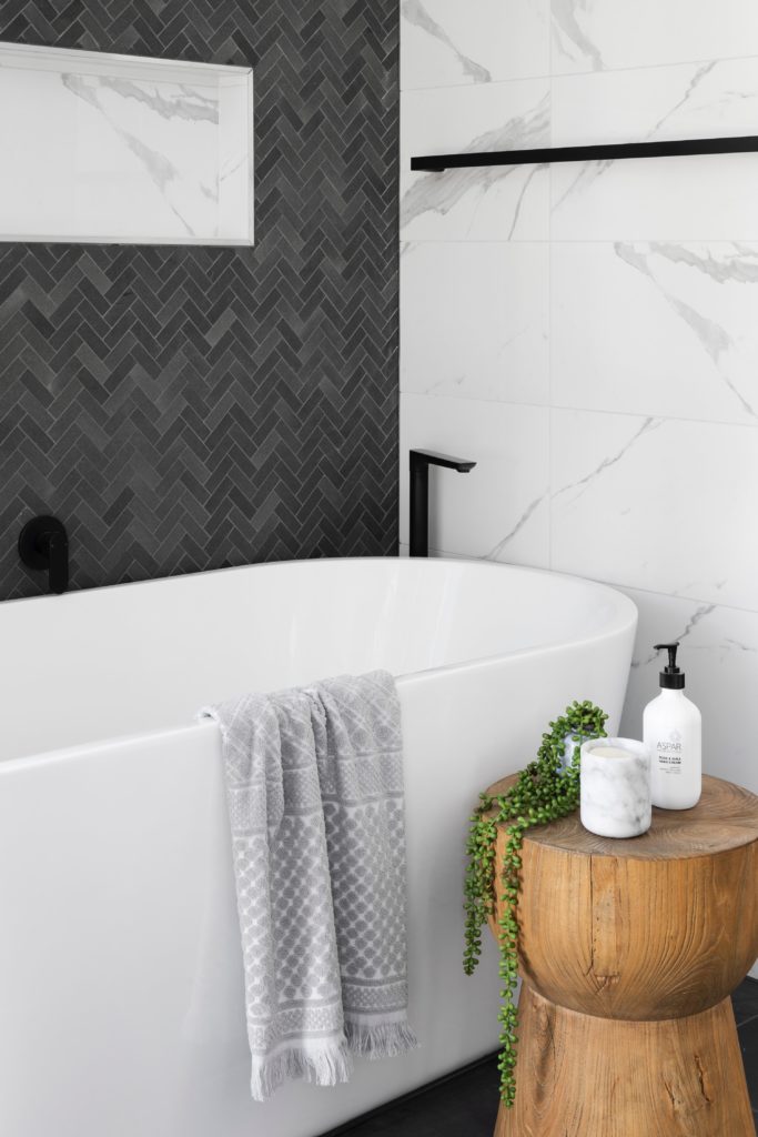 Accessorizing the bathroom: white marble and grey tile behind a large, white tub, next to solid wood pedestal containing a plant, candle and bath product.