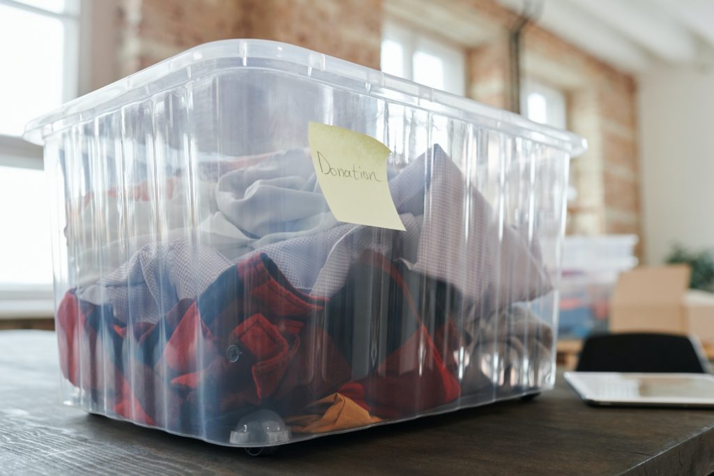 Minimalism and maximalism, part 2: a clear, plastic bin with clothing inside and a "donation" sticky note sits on a table in the foreground, a modern white and brick living room in the background.