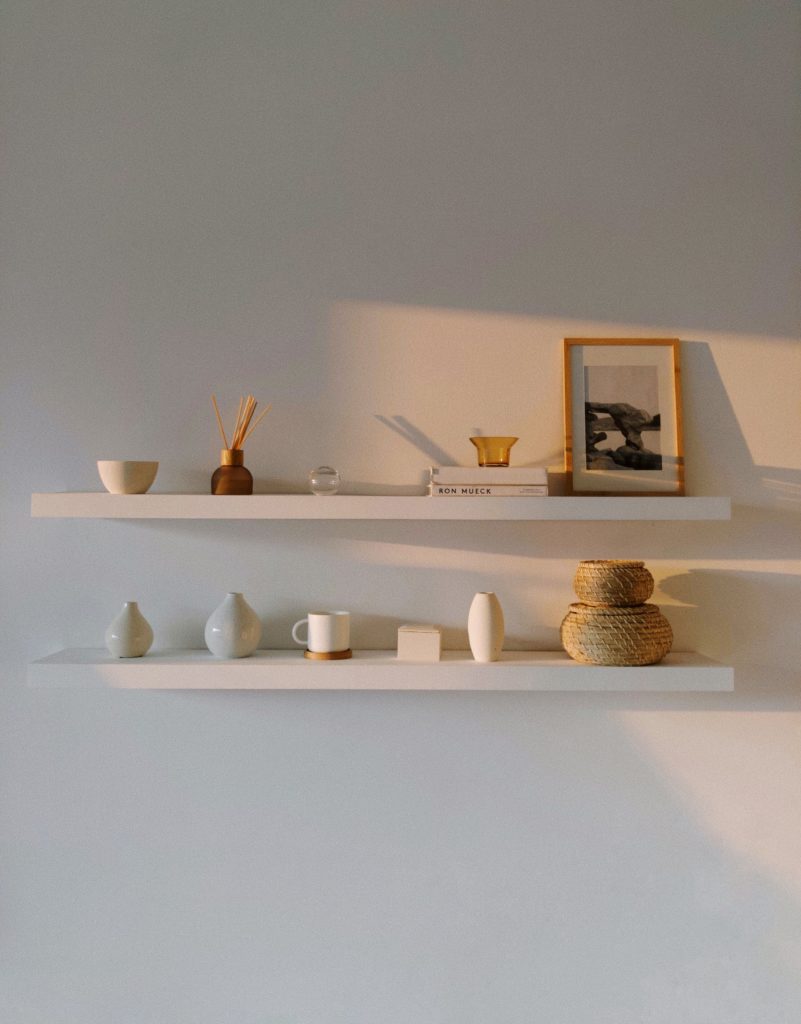 Minimalism and maximalism, part 2: a white wall with two white shelves that display small ceramic vessels and art, bathed in late day sun.