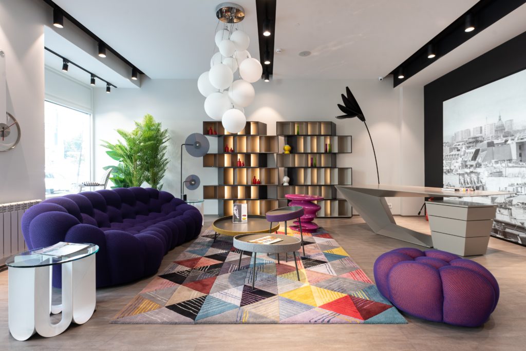 A modern and colorful living room, featuring organic shapes in sofa and ottoman, a muliticolored geometric rug, and asymmetric shelving.